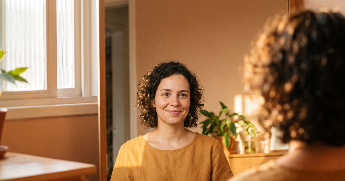 Mulher de blusa amarela sorri para o próprio reflexo no espelho, com a luz da janela ao lado